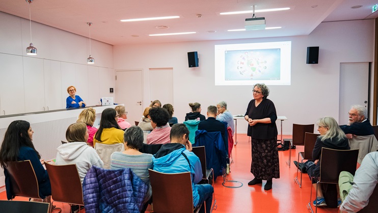 Saal mit Menschen in einer Stuhlreihe und Rednerin